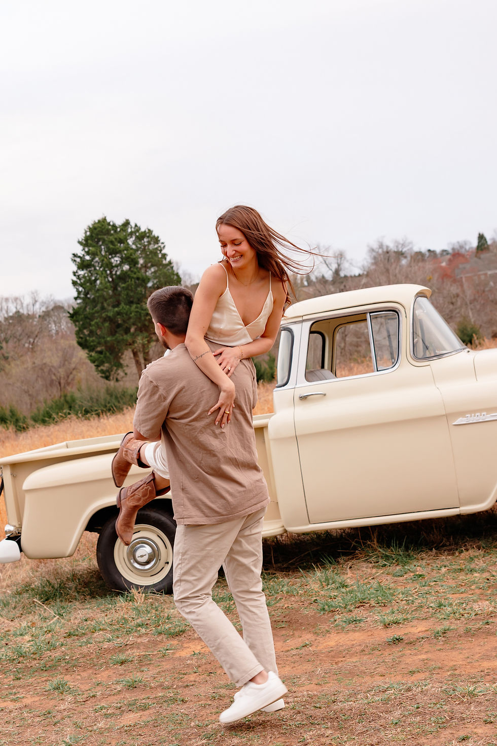 Man lifts smiling woman by a vintage beige truck in a grassy field; she's wearing cowboy boots, creating a joyful, playful mood. Vintage Chevy engagement shoot in Belmont, North Carolina