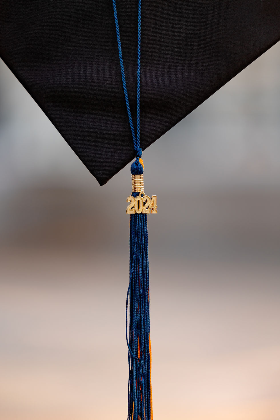 Graduation cap with a blue and gold tassel reading "2024" against a blurred background, symbolizing achievement and celebration. A woman in a white dress holds a graduation cap on a rooftop with blurred buildings in the background, during sunset. Senior cap and gown session in Charlotte, North Carolina.