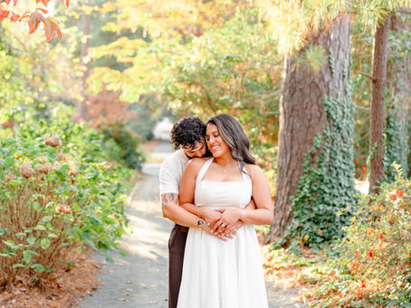 Couple embraces on a forest path in autumn. Woman smiles in white dress; man holds her lovingly. Vibrant foliage surrounds them. Charlotte Engagement Photos by Aubrey Elizabeth Photography