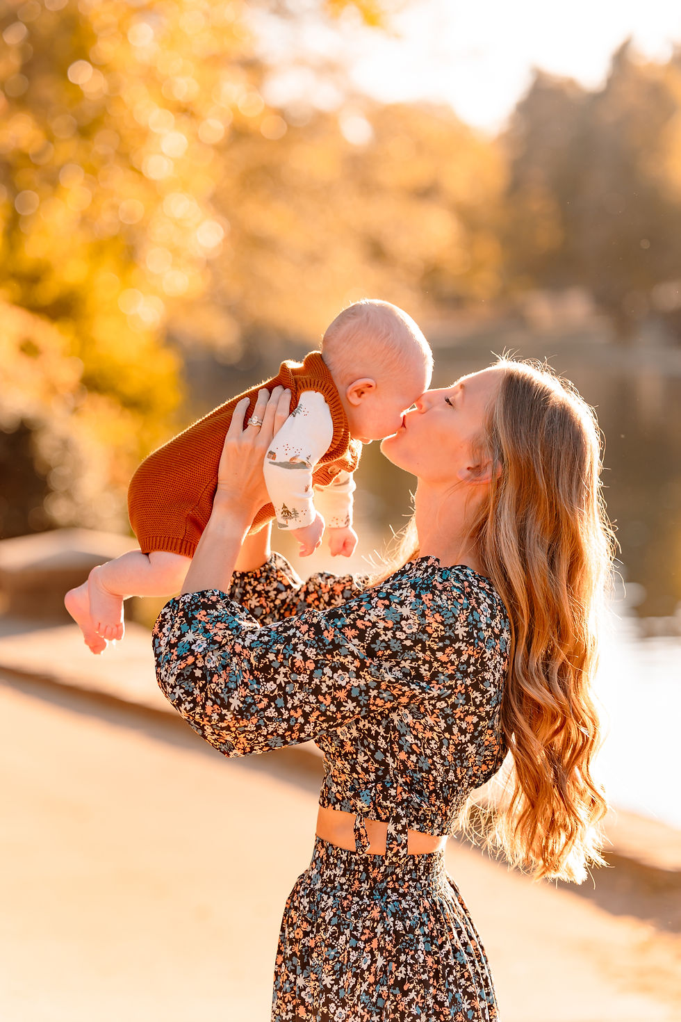 Woman in floral dress lifts baby in autumn park, both smiling. Baby wears brown outfit. Sunlit, golden background creates warm mood. Freedom Park, Charlotte, North Carolina.