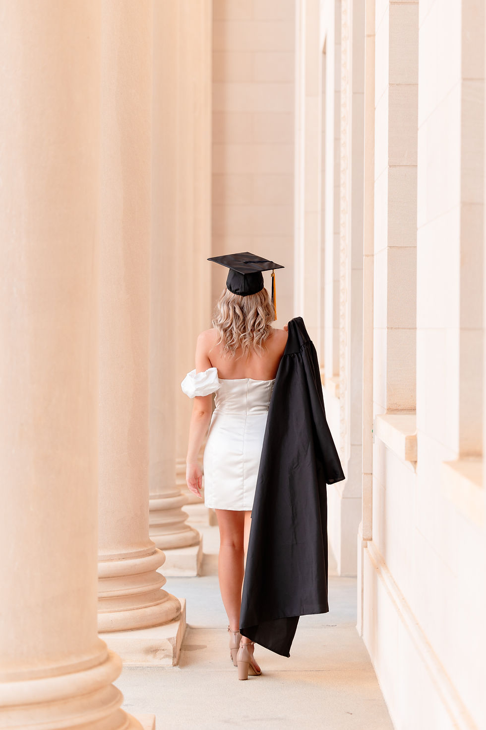 Graduate in a cap and white dress walks between stone columns, holding a gown over the shoulder. Neutral tones create a serene mood. A woman in a white dress holds a graduation cap on a rooftop with blurred buildings in the background, during sunset. Senior cap and gown session in Charlotte, North Carolina.