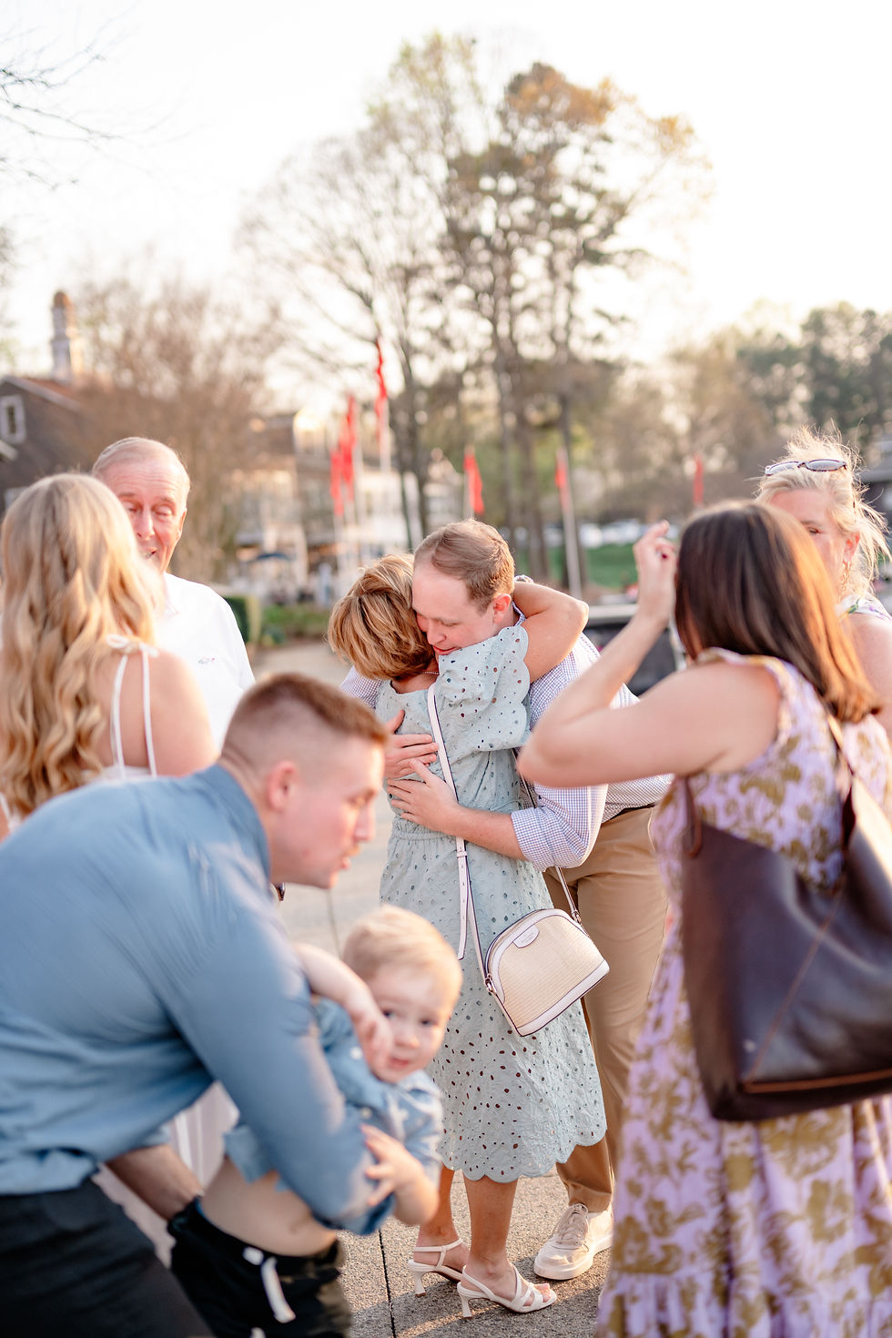 Group gathers outdoors, hugging and chatting. Trees in background, people in light dresses and shirts. Warm, joyful atmosphere. A man kneels proposing to a smiling woman in a white dress by a marina with boats. Trees and buildings in the background create a serene mood. Lake Norman Proposal at North Harbor Club.