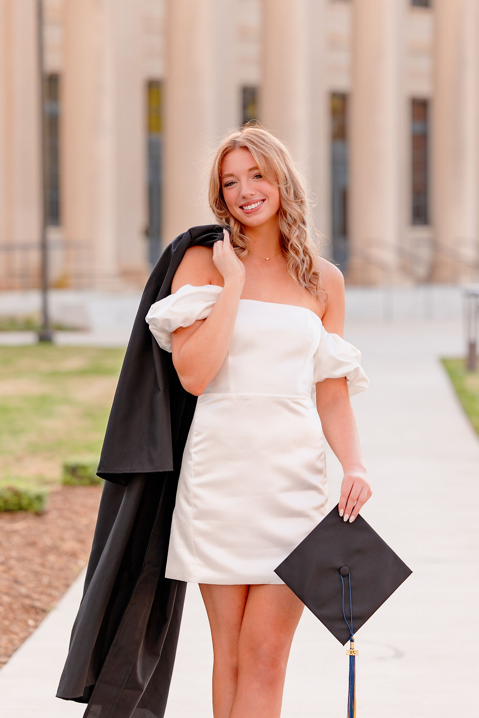 Young graduate in a white dress holds a cap and gown, smiling in front of stately columns, with a sunny, celebratory vibe. A woman in a white dress holds a graduation cap on a rooftop with blurred buildings in the background, during sunset. Senior cap and gown session in Charlotte, North Carolina.