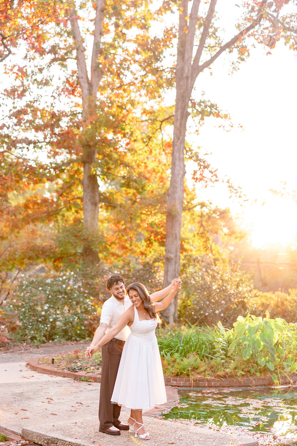A couple dances happily by a pond in a park with autumn leaves and sunlight. She wears a white dress; he wears a white shirt and brown pants. Couple embraces on a forest path in autumn. Woman smiles in white dress; man holds her lovingly. Vibrant foliage surrounds them. Charlotte Engagement Photos by Aubrey Elizabeth Photography