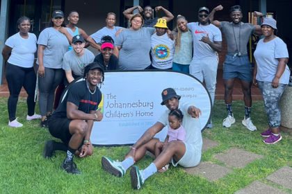 A group of volunteers and staff from Coaches For Charity and Lynx Professional Services posing with a banner for The Johannesburg Children's Home during a Stronger Together youth wellness and fitness outreach event.