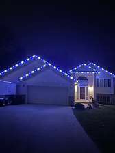 "Single-family home illuminated at night with professionally installed blue and white holiday lights outlining the roof peaks and entryway. The vibrant lighting creates a festive and polished look, showcasing AB Outdoor Solutions’ holiday décor service."