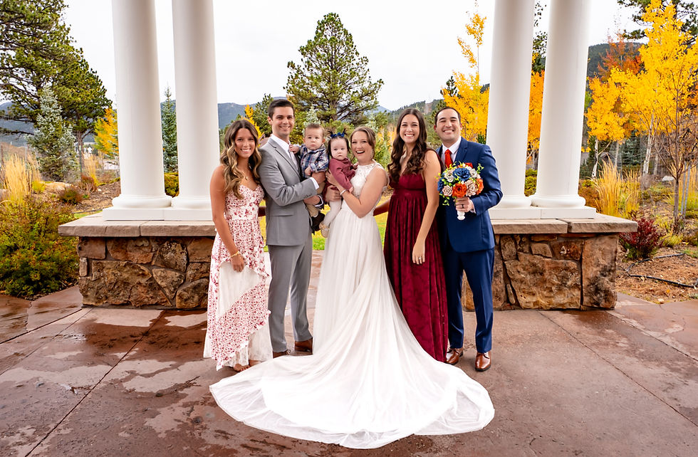 A group at an outdoor wedding poses by stone columns at The Stanley Hotel Pavilion in Estes Park, Colorado. Bride in white holds a baby, groom in gray holds another child. Autumn trees in background.