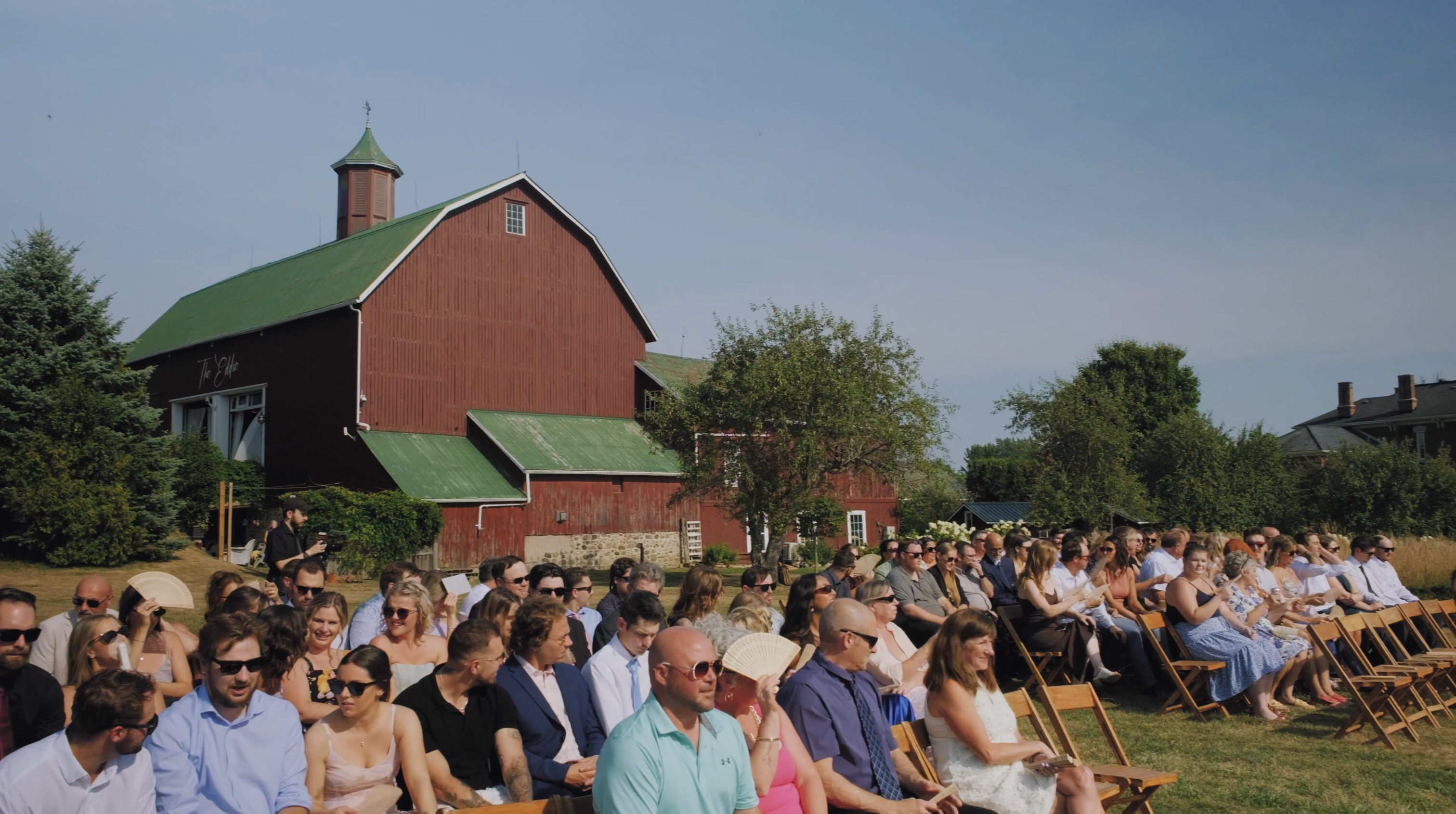 Outdoor wedding guests seated on wooden chairs by a red barn with green roof, dressed up and chatting under a clear blue sky