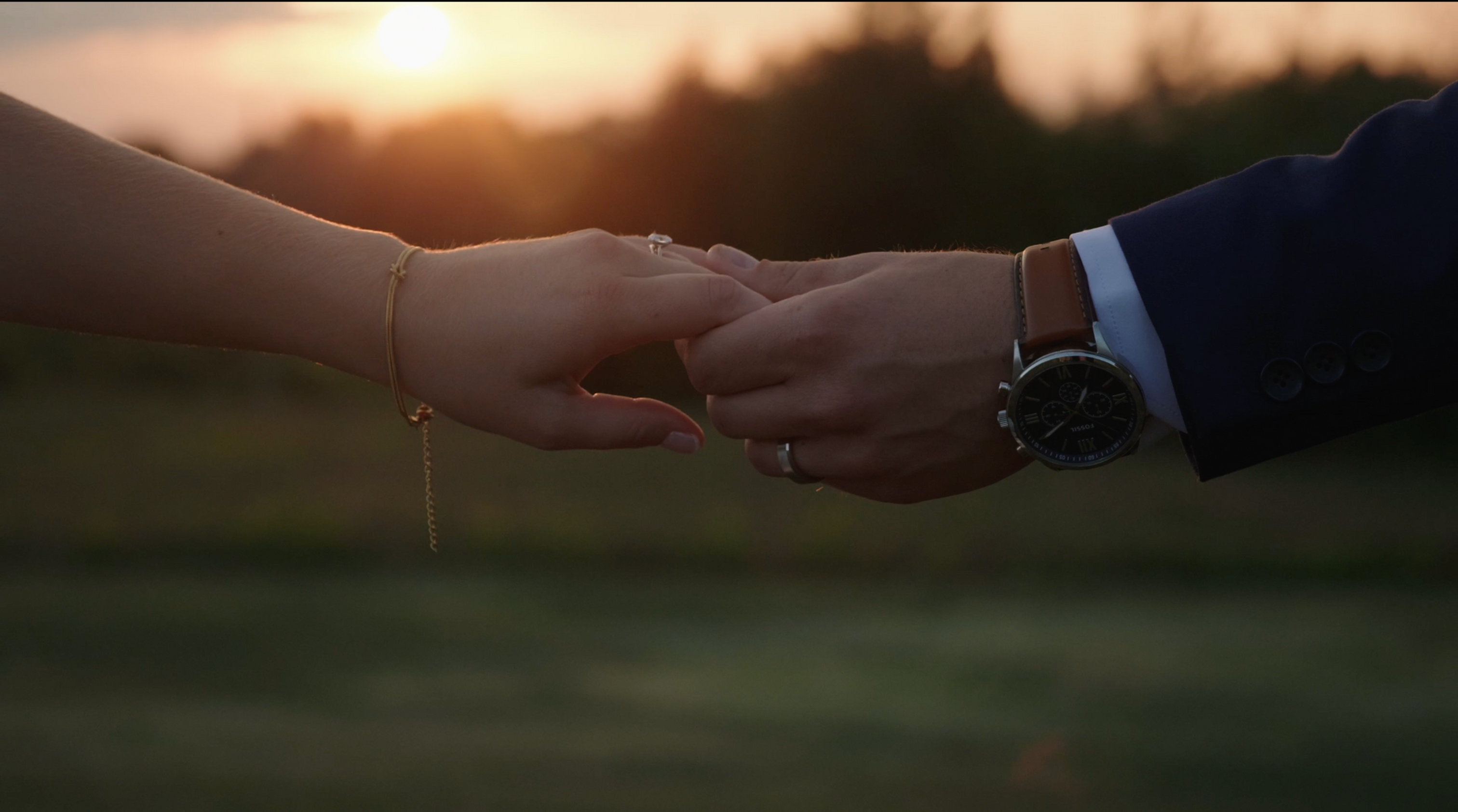 Couple holding hands at sunset, showing an engagement ring; man in suit with watch, warm romantic mood.