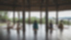 Five people meditate on mats in a wooden pavilion with a lush, green view outside. The scene is serene and peaceful.