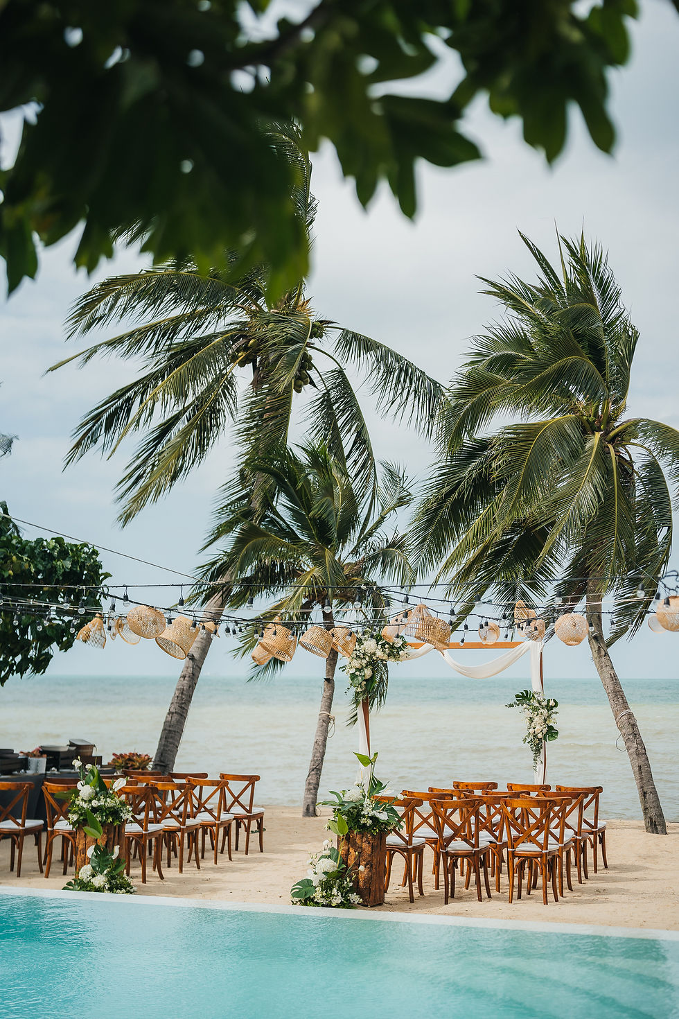 Beachfront wedding setup with palm trees, wooden chairs, floral arch and string lights beside a pool and calm ocean
