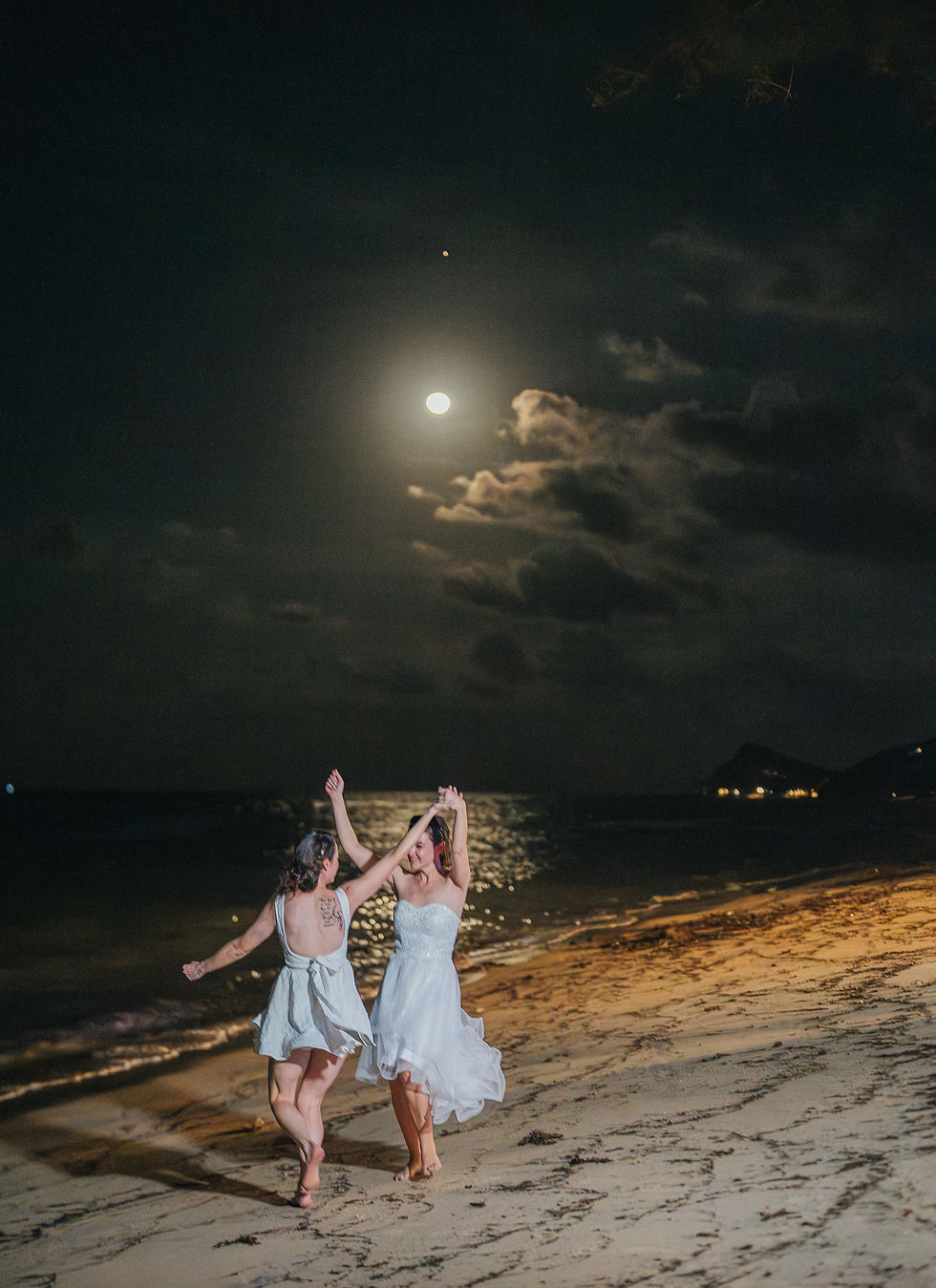 Two women in white dresses dance joyfully on a moonlit beach at night, with the ocean reflecting the full moon and scattered clouds above.