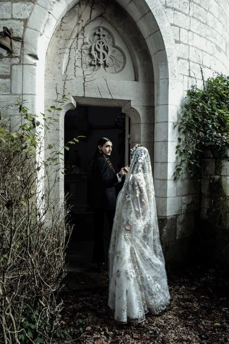 A person in a white veil stands outside a stone archway, holding hands with another person in black. Ivy-covered walls and a carved emblem above.