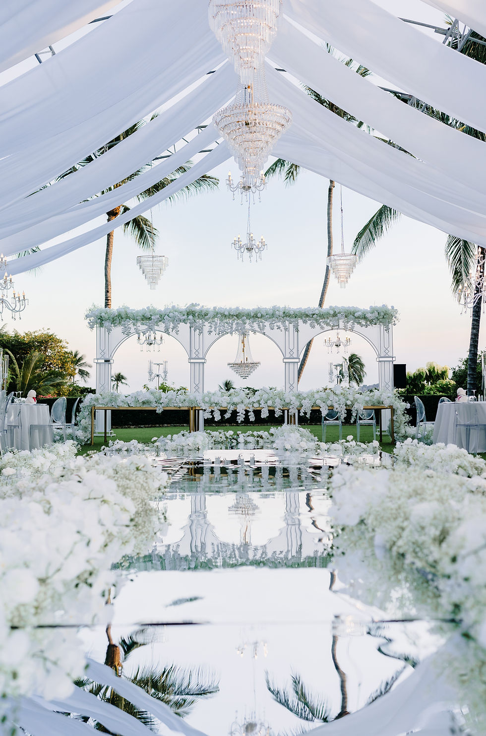Elegant outdoor wedding setup with white drapes, chandeliers, and flowers. Reflective path, palm trees, and clear sky in the background.