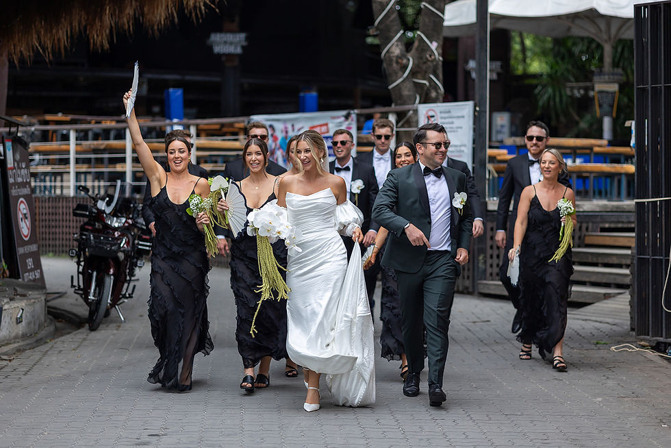 A bride in white and groom in dark suit joyfully lead a bridal party in black outfits down an urban street, holding flowers and fans.