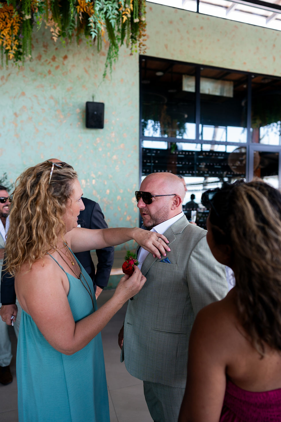 Woman pins boutonniere on man's suit in a tropical-themed setting. People around are dressed formally, with green and orange decor.
