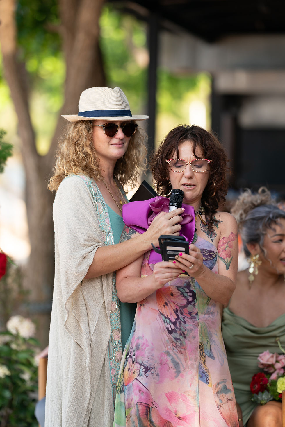 Two women outdoors, one in a hat and sunglasses, the other with a mic and phone. Colorful dresses and floral patterns. Relaxed setting.