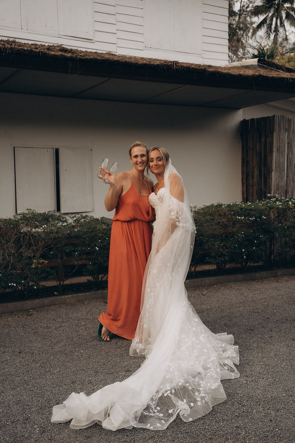 A Wedding Planner and her Bride smiling; one in a white wedding dress, the other in an orange dress holding champagne flutes. They're outside a white building.