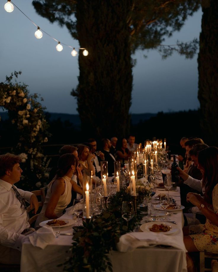 Dinner party outdoors at dusk, with elegantly dressed guests seated at a candlelit table under string lights, surrounded by trees.