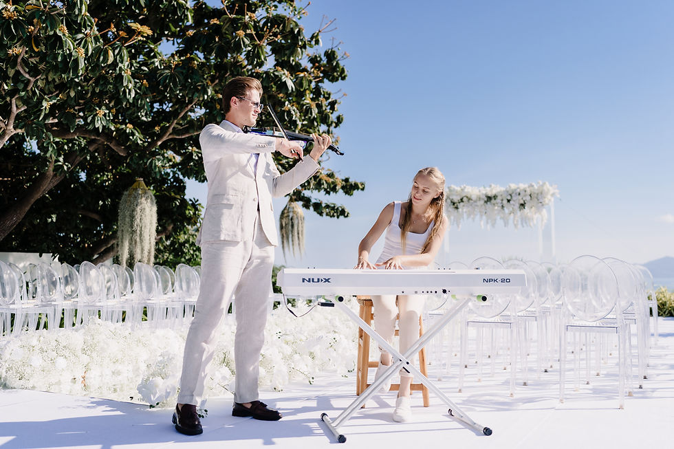 Man in white suit plays violin beside woman at NUX NPK-20 keyboard at an outdoor white floral wedding under a blue sky.