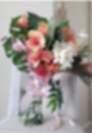 Elegant bouquet with pink, white, and orange flowers, lush green leaves, held by a hand. Set against a neutral, cushioned background.