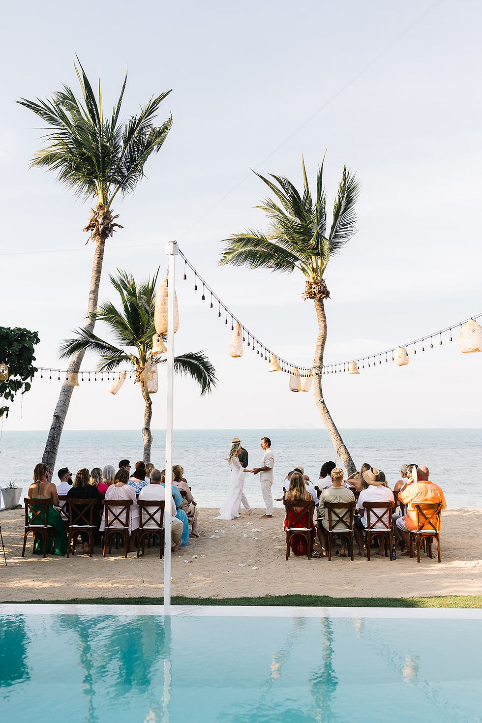 A beach wedding with a couple holding hands under palm trees. Guests sit facing the sea. Warm lighting, blue sky, and string lights create a serene mood.