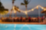Beachside dinner party at dusk with guests seated under glowing string lights and palm trees, reflected in a turquoise pool.