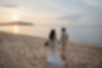 Bride in lace gown and groom in beige suit walk hand in hand on a serene beach at sunset, creating a romantic scene.