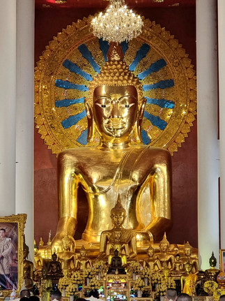Golden Buddha statue with ornate blue and gold halo, surrounded by smaller statues. White pillars and chandeliers adorn the temple interior.