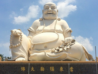Large, white laughing Buddha statue with a beaded necklace, sitting cross-legged against a blue sky. Chinese characters on the base.