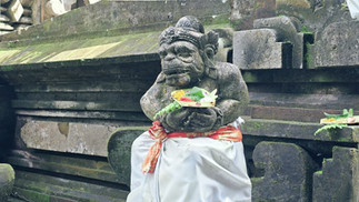 Moss-covered stone temple guardian statue with offerings, draped in white and red cloth, against dark carved stone walls.