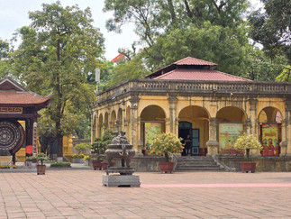 Historic yellow building with arches, surrounded by green trees and lanterns. Red text on wall: BẢO VẬT. Peaceful atmosphere.