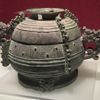 Ancient Chinese bronze vessel with intricate patterns and dragon handles on display against a red backdrop. Elegant and ornate design.
