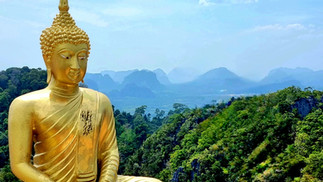 Golden Buddha statue sitting peacefully with lush green hills and misty mountains in the background under a clear blue sky.