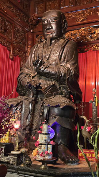 Bronze statue of a seated figure with intricate carvings, surrounded by ornate decorations, red lighting, and pink flowers in a wooden setting.
