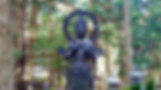 A dark statue of a serene figure stands in a lush forest. The background has tall trees and stone lanterns, creating a peaceful atmosphere.