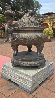 Ornate bronze incense burner with dragon designs on a pedestal, in a courtyard with trees and a historic building in the background.
