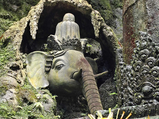 Carved elephant head with ornate patterns and a deity figure in a lush, moss-covered stone cave. Sunlight highlights intricate details.
