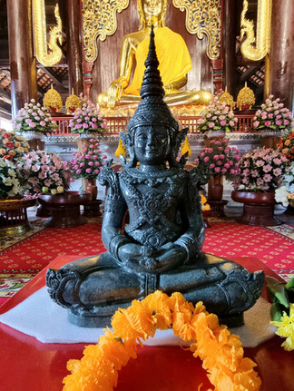 A black statue sits before a large golden Buddha adorned with flowers in a richly decorated temple. Yellow garland placed in front.
