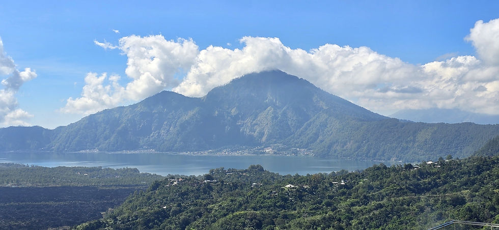 Wide lake below lush green volcanic mountains under bright blue sky with large white clouds, a calm scenic tropical vista