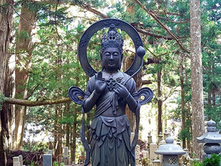 A serene statue of a deity stands amidst tall trees, surrounded by lanterns. The forest setting creates a peaceful atmosphere.