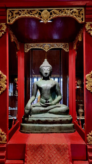 Seated Buddha statue in a red and gold ornate shrine, serene expression, with intricate patterns in a richly decorated room.