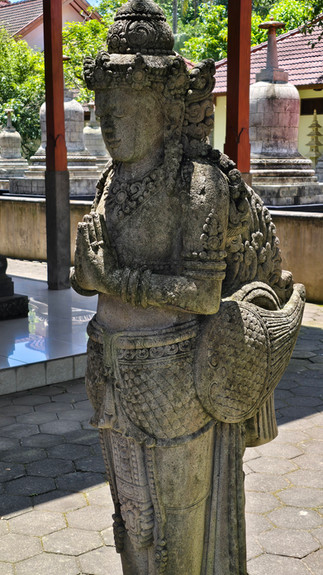 Stone statue of a serene figure with folded hands in a temple garden. Background of trees, tiled roofs, and other stone structures.