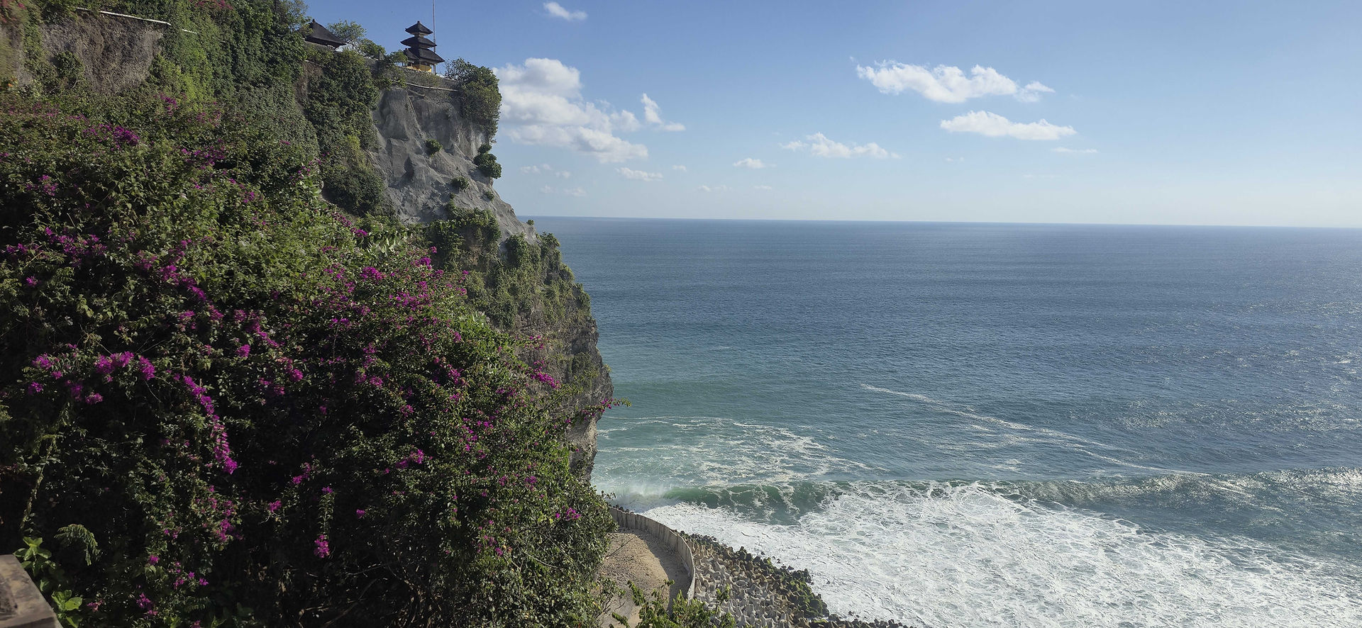 Cliffside temple surrounded by lush greenery and purple flowers, overlooking a vast, sparkling ocean under a clear blue sky.