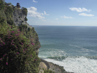 Cliffside temple surrounded by lush greenery and purple flowers, overlooking a vast, sparkling ocean under a clear blue sky.