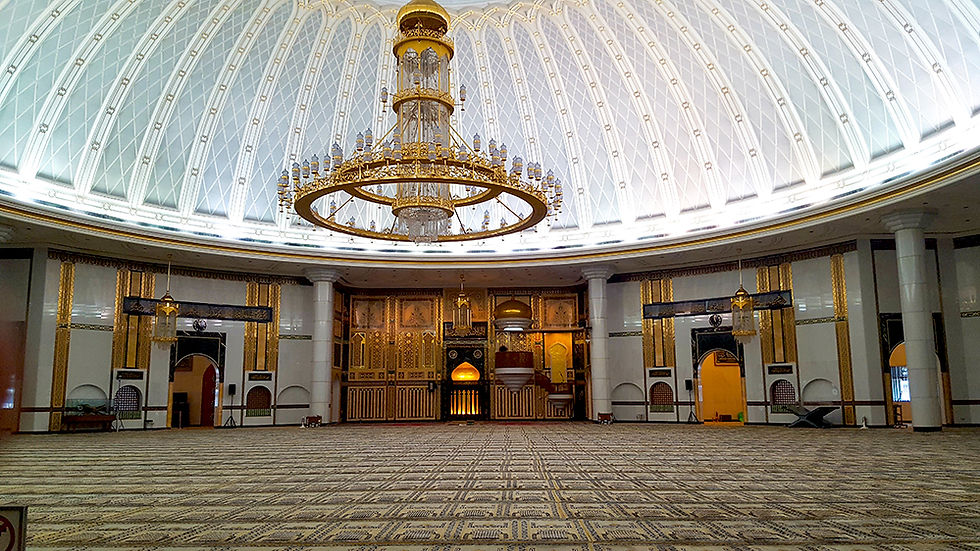 Spacious mosque interior with a large ornate chandelier, intricate gold and white decor, and a patterned carpet. Elegant, serene atmosphere.