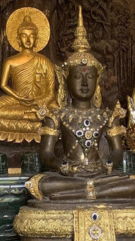 A dark statue adorned with gems sits cross-legged. A golden Buddha with a halo is in the background. Intricate carvings surround them.