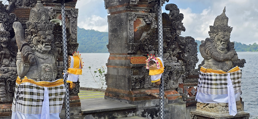 Carved stone guardian statues draped in yellow-white cloth stand at a lakeside temple, with a small warning sign and cloudy hills.