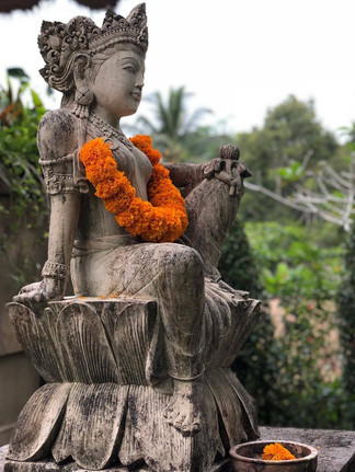 Stone statue of a seated deity adorned with bright orange marigold garland. Lush green background, serene and peaceful atmosphere.