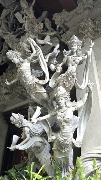 Intricate white stone dancer statues with ornate crowns and flowing scarves, stacked on a temple facade above green plants.